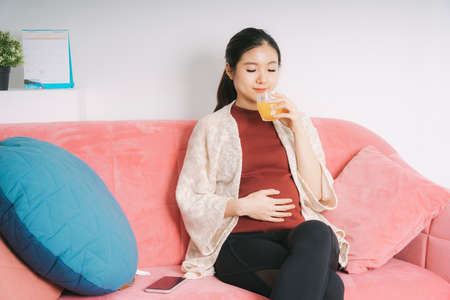 Beautiful pregnant asian woman drinking orange juice sit on a couch at living room.の写真素材