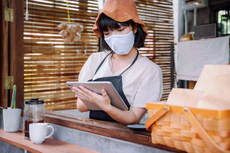 Beautiful asian thai woman wearing mask reopen coffee shop after covid-19 lockdown the city. Local business. Woman take order with digital tablet.の写真素材