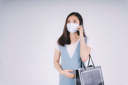 Beautiful pregnant asian woman wearing mask talking on the phone holding shopping back isolate over white background.の写真素材