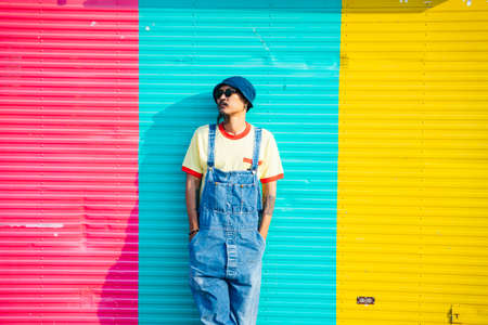 The man in yellow t-shirt and denim overalls standing and sitting in front of colorful shutter door in sunlight.の写真素材