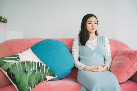 Young beautiful pregnant asian woman resting on a pink couch in living room.
