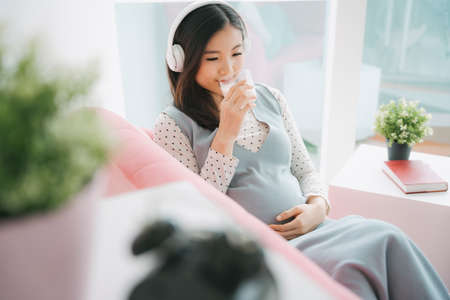 Young beautiful pregnant asian woman listen to musich with wireless headphone and drinking milk at home.の写真素材