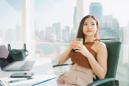 Portrait of Beautiful asian thai businesswoman sit on a chair holding healthy orange juice.の写真素材