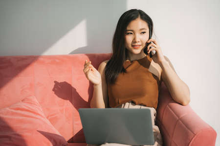 Beautiful asian thai businesswoman working on laptop and smartphone, sitting on a couch at home. Work from home.の写真素材