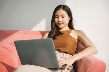 Beautiful asian thai businesswoman working on laptop at home on sofa. Work from home.の写真素材