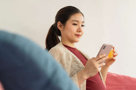 Beautiful pregnant asian woman holding orange juice and using smartphone sitting on a couch.
