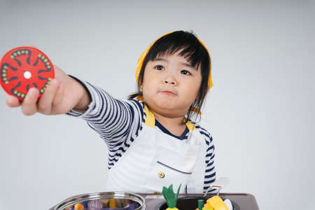 Pretty asian thai kid play role as a chef wearing yellow bandage holding slice of tomato cooking in kitchen.の写真素材