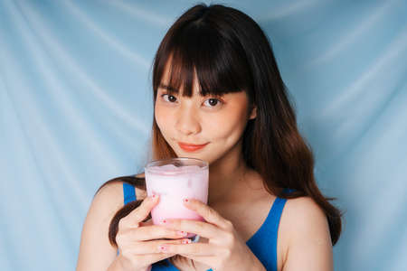 Cheerful young asian dark hair woman holding glass of pink milk shake with both hands.