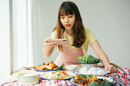 Portrait of young asian woman taking picture of Isan food with smartphone at home.の写真素材