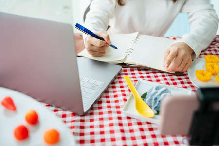 Food reviewer taking note on a book and searching for information on computer laptop.