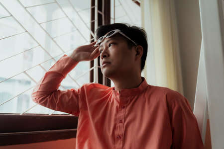 Asian gay in orange mandarin collar sitting at the window and raise his white eyeglasses up.の写真素材