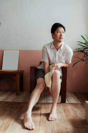 Asian black hair gay sitting on the wooden chair with white hat looking outside the window.の写真素材