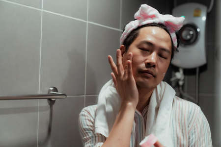 Black hair gay wearing pink headband with white towel around his neck and apply facial cream in the bathroom.の写真素材