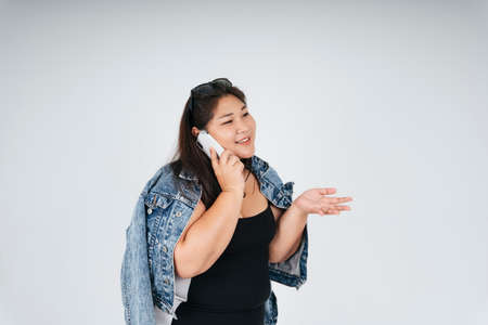 Portrait of cheerful happy asian chubby woman in black dress and jeans jacket talking on the phone on white background.の写真素材