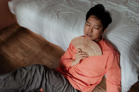 Black hair gay in orange mandarin collar sit on the wooden floor lean at the white bed hold the...