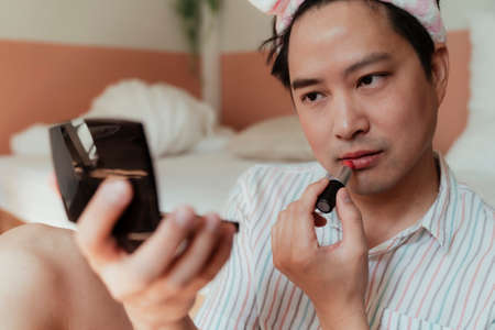 Asian gay with pink headband looking at the small mirror while apply red lipstick on his lip.の写真素材