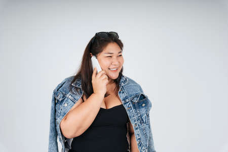 Portrait of cheerful happy asian chubby woman in black dress and jeans jacket talking on the phone on white background.の写真素材