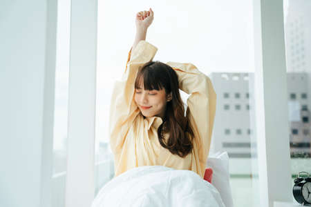 Portrait of young asian dark hair woman wake up on bed in the morning. Girl stretching arms relax after woke up.の写真素材