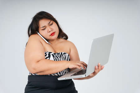 Busy asian chubby woman holding laptop and talking on the phone with serious looking on white background.の写真素材