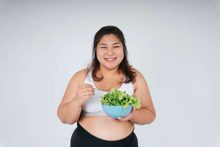 Cheerful happy asian chubby woman in white sport bra enjoy eating salad bowl over white background.の写真素材
