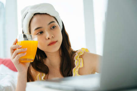 Young asian dark hair woman holding glass of orange juice using laptop computer.