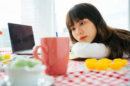 Young asian dark hair woman looking at thai dessert on table while working with laptop.の写真素材