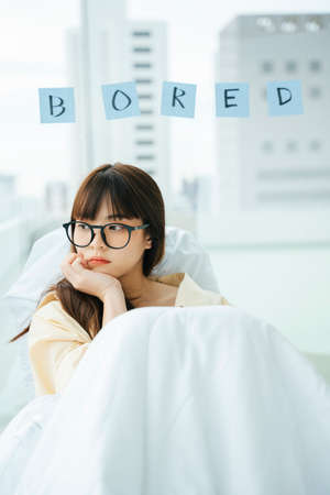 Cute young asian dark hair woman wearing eyeglasses laying on a bed. Bored girl relax on a couch.の写真素材