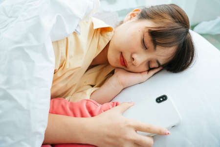 Young asian dark hair woman sleeping on a bed while holding smartphone.の写真素材