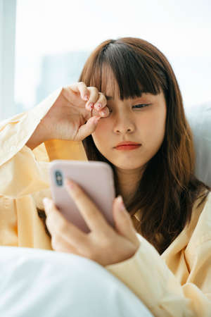 Sleepy young asian dark hair woman wake up on a bed and use smartphone in the morning.の写真素材
