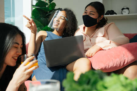Diversity society concept. Asian and african women wear face shield and mask using laptop at home.