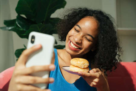 African curly hairstyle woman doing live streaming on smartphone about eating donut. Food vlogger.