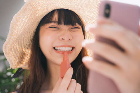 Close up portrait of smile asian traveler woman wearing hat applying lipstick and looking at mirror on smartphone.の写真素材