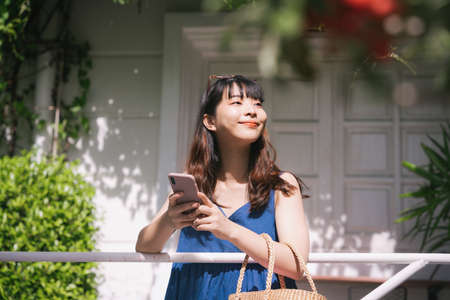 Cheerful happy young asian woman traveler wearing blue dress booking hotel or order stuff with online service with smartphone.の写真素材