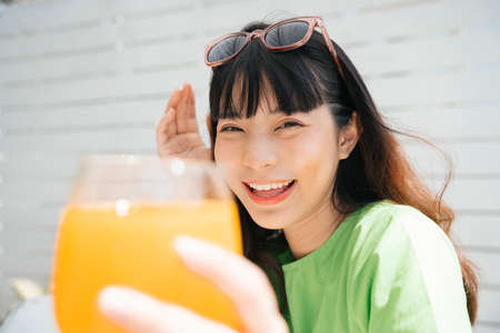 Cheerful young asian woman traveler wearing green shirt and sunglasses enjoy drinking healthy orange juice.の写真素材