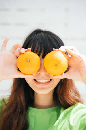 Young asian woman traveler wearing green shirt holding orange fruits at eyes.