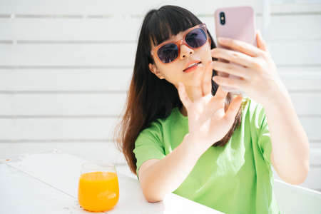 Beautiful young asian woman traveler wearing sunglasses holding glass of orange juice doing vlog with smartphone for social media.の写真素材