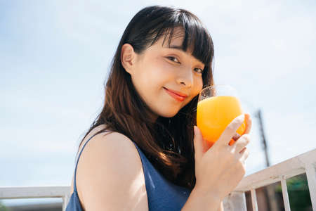 Young asian woman traveler wearing blue dress enjoy drinking healthy orange juice outdoors at balcony.の写真素材