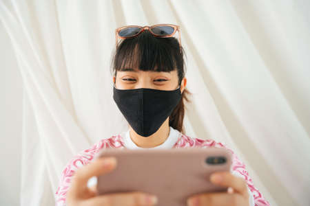 Young asian woman in pink shirt wear mask and sunglasses enjoy shopping or playing game on smartphone.の写真素材
