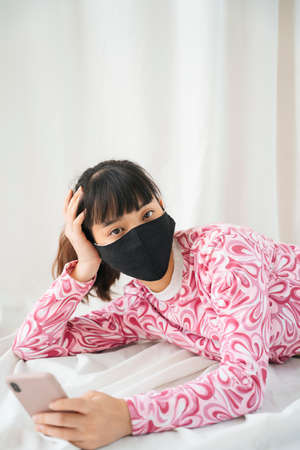 Young asian woman in pink shirt wearing mask laying on a bed, using smartphone.の写真素材