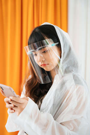 Young asian woman wearing white raincoat and face shield using smartphone.の写真素材