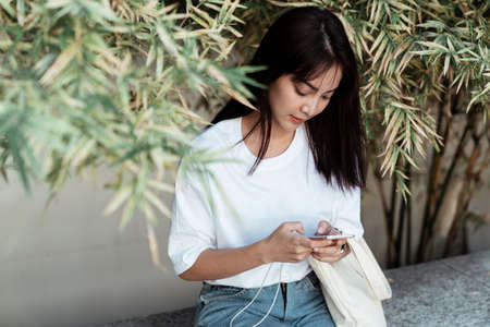 Cute girl in white t-shirt sitting at the little bamboo garden and using the phone.