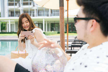 Beautiful asian woman taking photo of her boyfriend with smartphone at hotel swimming pool.