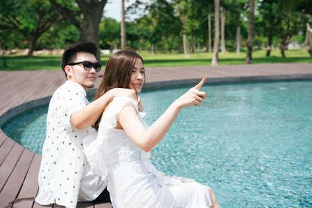 Asian couple traveler sitting on the edge of swimming pool together. Woman pointing out at something.の写真素材