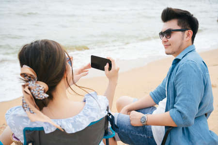Asian couple traveler sitting on chair taking photo with smartphone on the beachの写真素材