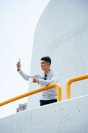 Portrait of stylish asian adult man taking selfie with coffee cup at balcony.
