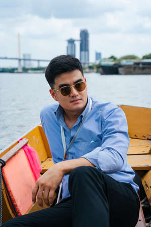 Fashion portrait - Asian business man wear sunglasses sitting on wooden boat in Bangkok.