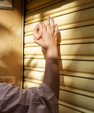 Cropped image of fat woman's hand raise pink donut at the yellow shutter door with sunlight.