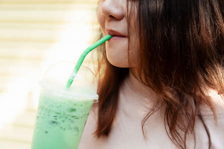 Cropped image of iced green tea in plastic cup is drunk with green straw by long hair fat woman.の写真素材