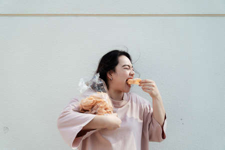 Long hair fat woman eat rice craker with wide mouth in front of white wall.の写真素材
