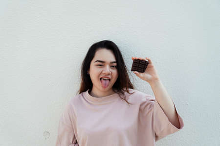 Long hair fat woman standing at the white wall with chocolate bar in her hand.